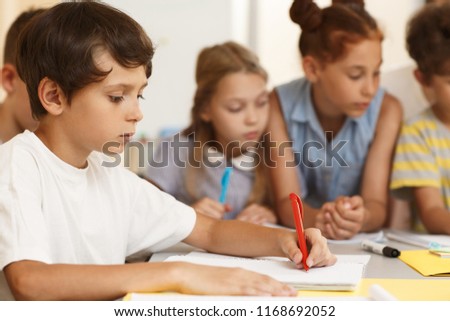 Side view of serious little student sitting at table and writing essay in classroom. Hardworking pupil in white shirt doing test while classmates studying material in team. Concept of learning. Royalty-Free Stock Photo #1168692052