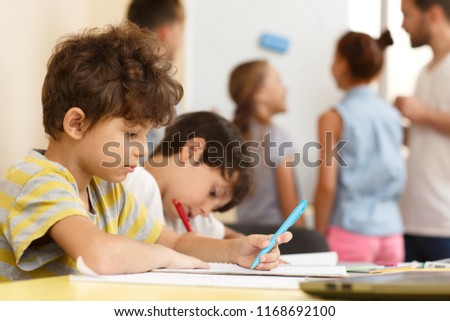 Side view of hardworking schoolboy carefully doing tasks in copybook during lesson on interesting courses. Cute little boy sitting at table and writing. Concept of modern education and learning. Royalty-Free Stock Photo #1168692100