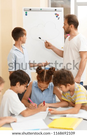 Stylish schoolboy with classmates sitting and working at table in process of learning in modern school. Pupil and male teacher standing near magnetic board and discussing questions.
