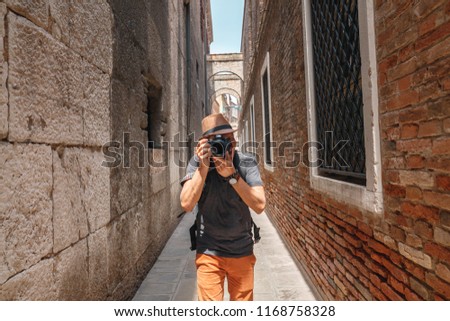 Young man in hat with backpack taking photo with his camera. Man with professional camera looking through lens