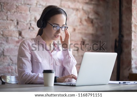 Young concentrate female in eyeglasses wearing headphones using  computer in office for online study, self-tuition, learning foreign languages, listening music, working assistance in internet concept