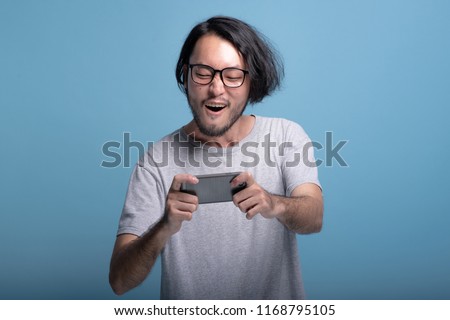 Excited young bearded man playing mobile games in blue background. Happy Asian young hipster playing mobile game, half body shot. Young generation hipster relaxing concept. Royalty-Free Stock Photo #1168795105