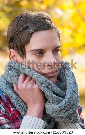 Attractive short haired pensive woman snuggling into a warm knitted winter scarf to keep out the autumn chill with colourful autumn foliage in the background