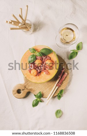 Melon and ham or prosciutto salad served in half of Cantaloupe melon, decorated fresh basil and grissini bread on wooden serving board over white linen cloth with glass of white wine. Flat lay, space