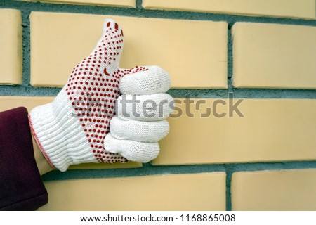 Home improvement.A gesture of approval. The hand of a man in a glove with his finger raised up against a yellow brick wall.