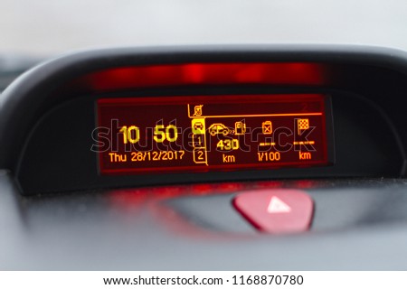 Digital lcd screen of a car onboard computer showing date and time, fuel consumption info and trip lenght in kilometers. Also hazard warning lights button visible on foreground. Royalty-Free Stock Photo #1168870780