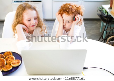 Brother and sister are sitting in the kitchen at the computer table watching cartoons