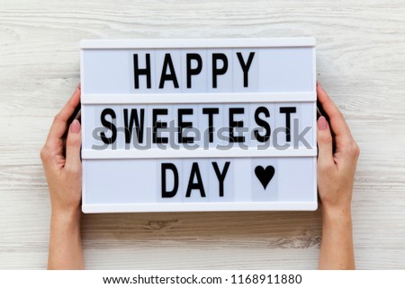 Female hands hold modern board with text 'Happy Sweetest Day' word over white wooden background, top view. From above, flat lay, overhead.  Royalty-Free Stock Photo #1168911880