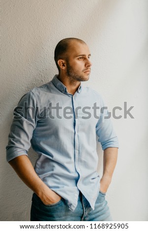 Brutal man in the blue shirt and blue jeans shorts posing holding hands in pockets in the the flat with white walls near the window in Spain
