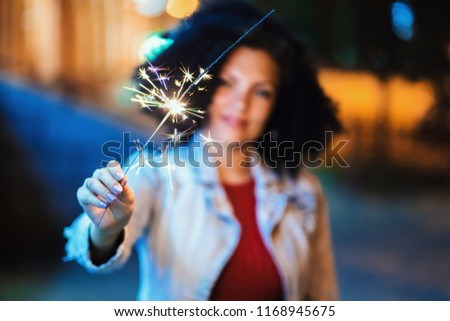 Young beautiful woman with very curly afro hair dancing with bengal fire at night illuminated street. Unusual trendy girl with sparklers. Holiday concept.