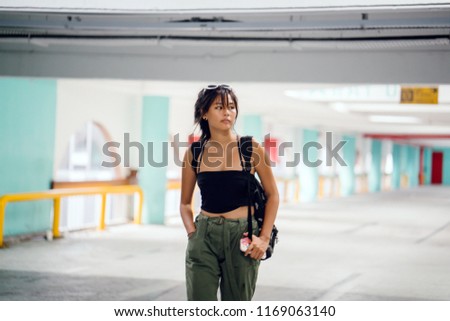 Portrait of a cool, young and attractive millennial teenager girl in trendy street clothing, sneaker and a backpack. She is posing for her photo in a gritty car park in the daytime.