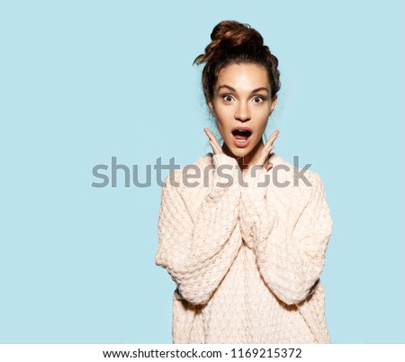 Portrait of surprised beautiful woman holding hands near face and looking at camera with surprised on blue background. Brunette model dressed in knitted pullover. Autumn fashion concept. 