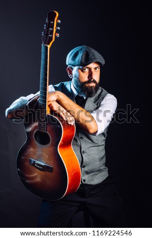 A bearded man in suit and hat sitting and playing on the acoustic guitar. Studio portrait
