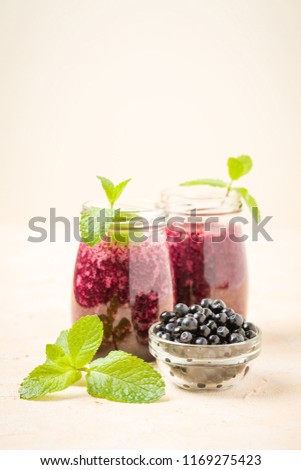 Blueberry smoothie decorated with fresh green mint leaves and raw ripe berries on yellow pastel background - transparent jars of summer blended vitamin cocktail with copy space.