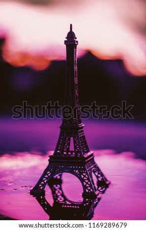 A toy eiffel tower stands in a puddle reflecting the purple sunset.