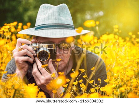 Photographer in white hat with vintage style camera in beautiful summer meadow of yellow buttercups taking a picture.