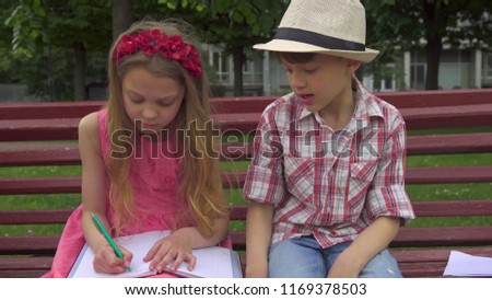 Pretty little girl girl drawing in her album on the becnh. Handsome caucasian boy in hat telling something his female friend. Cute kids holding pens in their hands