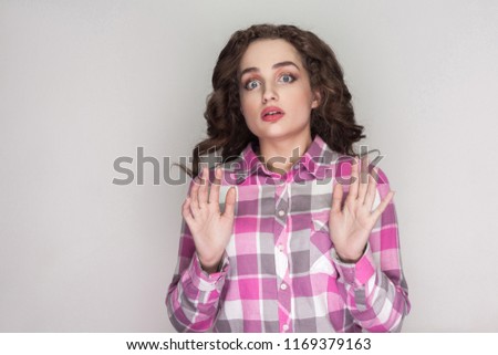 scared or shocked girl with pink checkered shirt, curly hairstyle and makeup standing, holding hands up and looking at camera with confused face. indoor studio shot, isolated on gray background.
