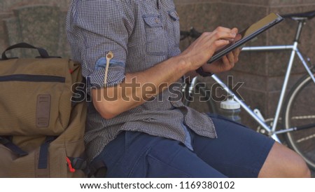 Male cyclist flipping on tablet touchscreen on the street of the city. Low shot of sporty guy sitting on the edge against background of his bike. Close up of young man using some digital gadget near