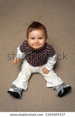 A cute baby girl sits on the floor looking up at the camera.  She is wearing Ponseti Method Shoes for Club Foot adjustment the bar is not clicked in in the picture so she can practice weight bearing.