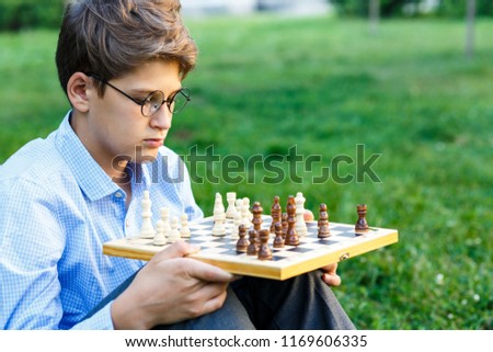 cute young boy in blue shirt holds chess board  and plays chess in the summer park. Education, intellectual game 