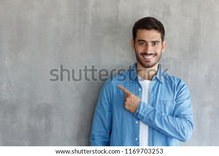Young handsome man in blue shirt pointing left with his finger, standing against on gray textured wall