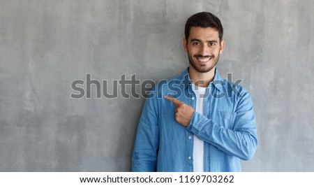 Attractive young man in blue shirt pointing left with his finger isolated on gray textured wall