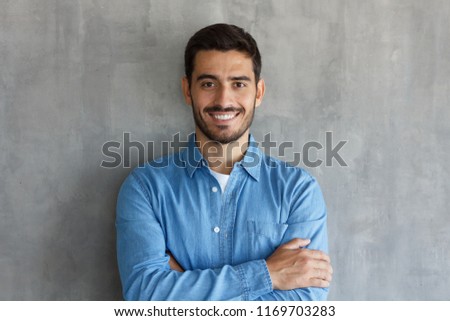 Smiling man in blue shirt, standing with crossed arms against gray textured wall 