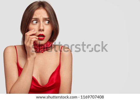 Image of scared frightened nervous beautiful young lady looks aside, keeps hand near opened mouth, dressed in red clothes, stands against white background with copy space for your promotion.