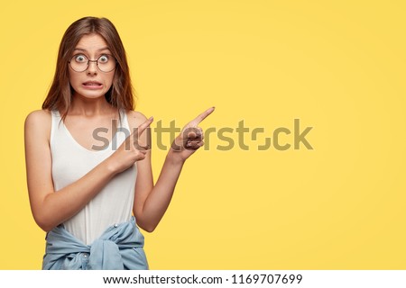 Oops, come and look there! Lovely young woman feels worried and ashamed, points with both index fingers at upper right corner, isolated over yellow background, makes mistake and looks awkward Royalty-Free Stock Photo #1169707699