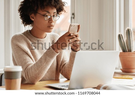 Cropped image of serious beautiful dark skinned freelancer holds smart phone, recieves new task, reads notification with attentive look, works on laptop computer at home, sits in workplace alone Royalty-Free Stock Photo #1169708044