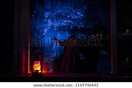 Horror Halloween concept. Burning old oil lamp standing on window with silhouette of an unknown shadow figure with backlight. Night scenery of a nightmare scene. Selective focus.