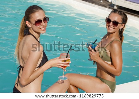 Two beautiful happy female friends smiling to the camera while having drinks by the swimming pool. Beautiful women enjoying their summer vacation, relaxing by the pool. Friendship, travelling concept