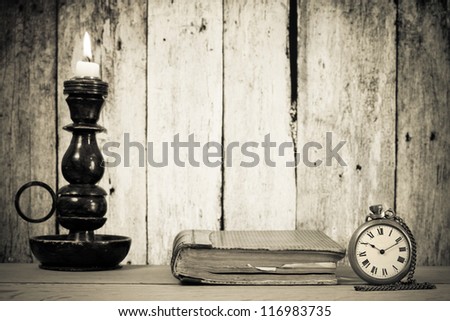 Vintage pocket watch, book and candlestick with burning candle on a table in front of wooden background