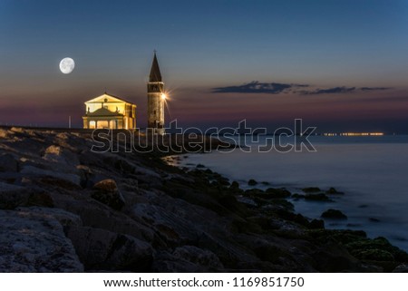 Enlightened church and tower "Santuario della Madonna dell'Angelo" in the town of Caorle on the coast at night with shining moon above the church. Royalty-Free Stock Photo #1169851750