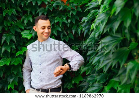 the guy waits for someone and looks at the watch against the background of the building on the facade of which is a plant with green foliage
