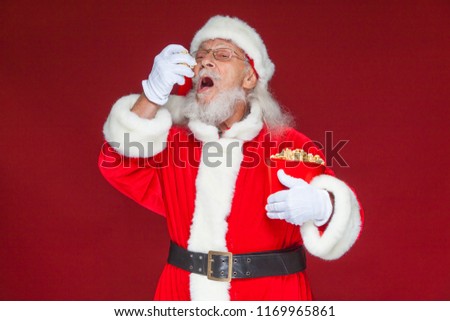 Christmas. Smiling, kind Santa Claus in white gloves with his mouth open holds a red bucket with popcorn with one hand, takes some popcorn and prepares to eat it with the second hand. The concept of