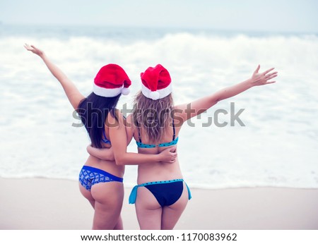 Back view of two beautiful girls in bikini in santa hat staying on the beach