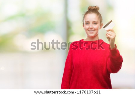 Young blonde woman holding credit card with a happy face standing and smiling with a confident smile showing teeth