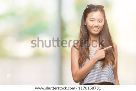 Young asian woman wearing sunglasses over isolated background cheerful with a smile of face pointing with hand and finger up to the side with happy and natural expression on face 
