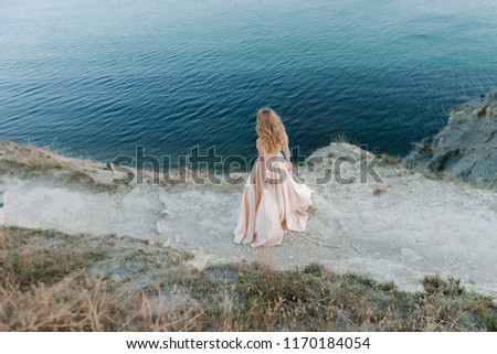 Beautiful blonde girl with a chic piling in a lush peach dress stands against the blue sea on the horizon at sunset