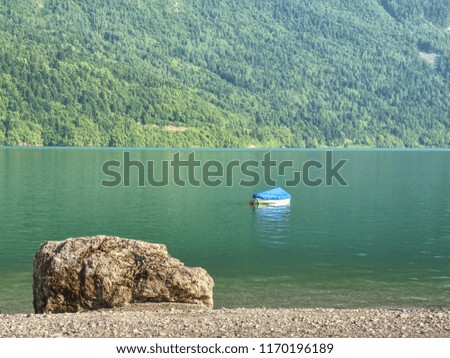 Boat on  green blue water level of Alps lake. White stony shore of a mountain lake. Reflection of forest mountains in water level.