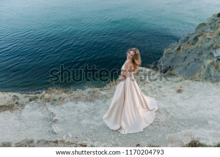 Beautiful blonde girl with a chic piling in a lush peach dress stands against the blue sea on the horizon at sunset