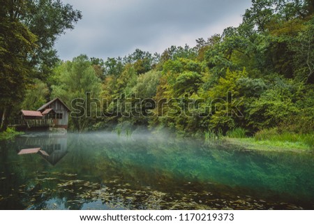 River source or spring of Krupa in Bela Krajina (White Carniola), Slovenia on a misty cloudy day. Reflection of wooden house, pier mill in foggy green river. Scary mystical water concept photo.