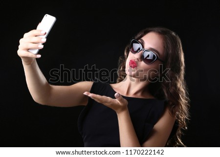 Young woman sending kiss and making selfie on black background
