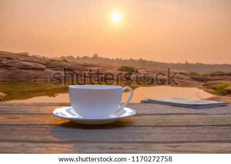 A selective focus picture of a cup of coffee on wooden floor in the morning sunrise beside lake.