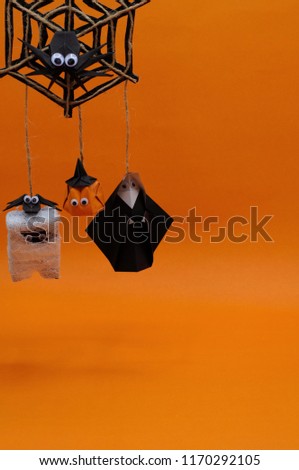 The origami Halloween background of pumpkin head jack-o-lantern, mummy and nun hanging on spider cobweb isolated on orange background with space for text.