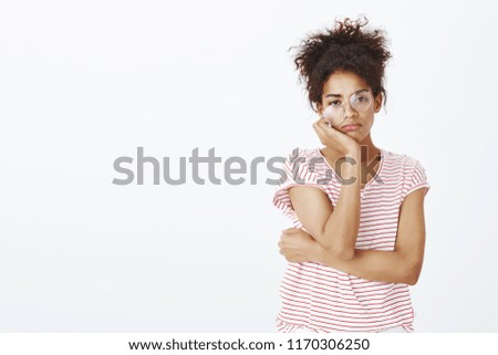 Studio shot of bored indifferent cute daughter in striped t-shirt and glasses, looking with careless expression at camera, leaning head on hands, being unimpressed and having no care over gray wall