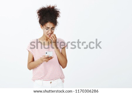 Indoor shot of nervous insecure dark-skinned woman in striped t-shirt and stylish eyewear, biting fingernail, frowning and looking at smartphone screen, being unsure while sending important message