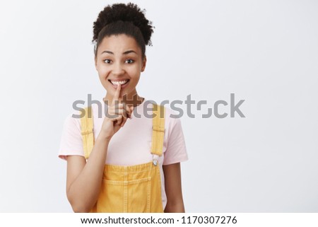 Keep secrets safe. Portrait of charming friendly-looking cute African American female in yellow trendy overalls, saying shh while showing shush gesture, smiling and holding index finger over mouth